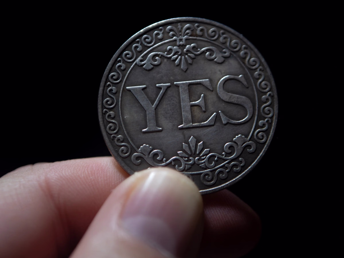 Engraved ornamental YES coin held between fingers — Source Select.